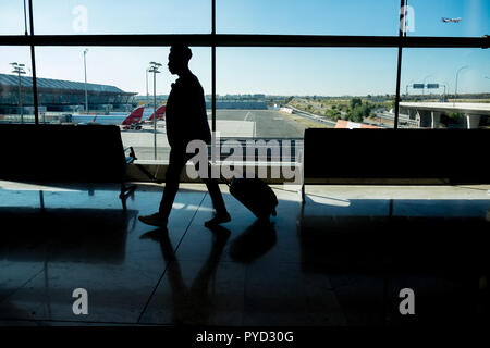 Un giovane imprenditore è camminare attraverso l'aeroporto portando una valigia di fronte alla finestra con gli aerei in attesa in le dita dietro il vetro Foto Stock