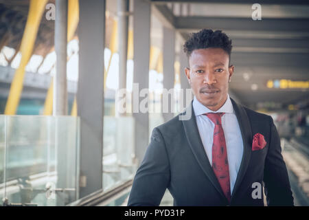 In prossimità di una giovane e attraente imprenditore nero guardando la telecamera mentre si è in attesa di una vettura al di fuori dell'aeroporto indossando un abito nero Foto Stock