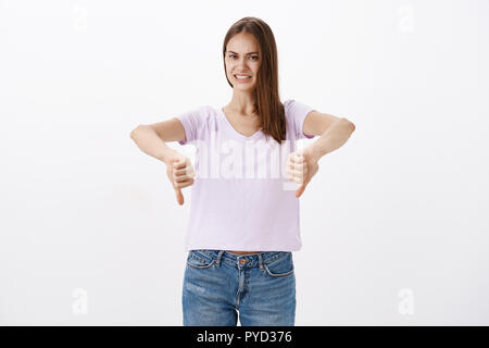 Studio di shto orinato insoddisfatto attraente giovane femmina cliente in camicetta e jeans che mostra il pollice verso il basso sorridendo goffamente offrendo al contempo un feedback negativo e di rifiuto cattiva idea oltre il muro bianco Foto Stock