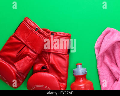 Cuoio rosso guantoni da pugilato, una bottiglia d'acqua in plastica e un asciugamano rosa su sfondo verde, lo spazio vuoto nella parte superiore Foto Stock