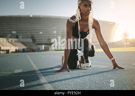 Atleta femminile di prendere posizione sul suo segna per iniziare la corsa. Guida femmina mettere in pratica il suo sprint su un all-weather via di corsa in uno stadio. Foto Stock