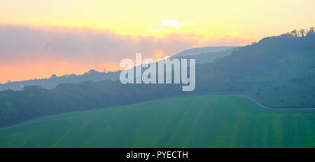 Campagna britannica che si affaccia su un ampio campo di verde a filari di alberi e di una collina è il sole che sorge dietro la collina e dando un bagliore arancione t Foto Stock