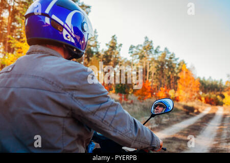Senior uomo scooter di equitazione in autunno la strada forestale. Driver nel casco ciclomotore di equitazione. Vista posteriore Foto Stock