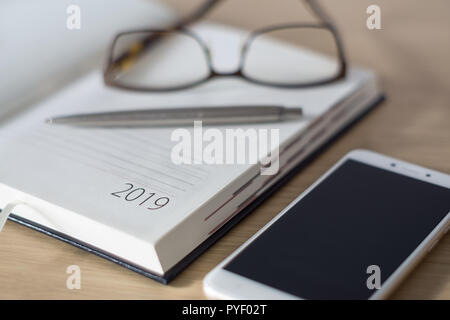 Nuovo anno 2019. Office organizer Calendario, smartphone, bicchieri e sliver penna a sfera sulla scrivania in legno. Messa a fuoco selettiva. Foto Stock