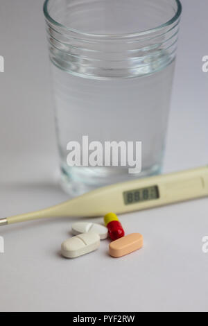 Pillole in diversi colori, un termometro e un bicchiere di acqua su sfondo bianco, medicina Foto Stock