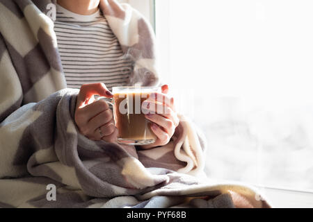 Cottura a vapore caldo bere nella donna con le mani in mano. Femmina coperto di gettare una coperta si siede da finestra con tazza di cioccolata calda Foto Stock