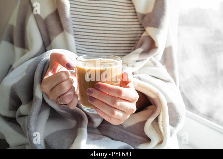 Cottura a vapore caldo bere nella donna con le mani in mano. Femmina coperto di gettare una coperta si siede da finestra con tazza di cioccolata calda Foto Stock