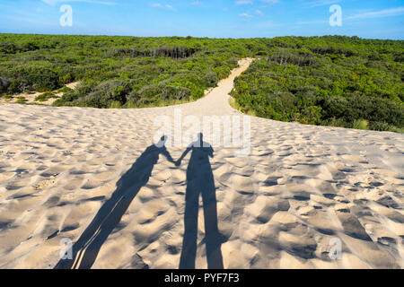 Ombre di un uomo e di una donna in piedi insieme nelle dune di sabbia al tramonto, vicino all'ingresso del cammino che conduce alla selvaggia foresta di pini . Foto Stock