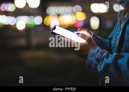 Donna puntare il dito sulla schermata vuota smartphone su sfondo bokeh luce nella notte città atmosferica, hipster utilizzando in mani pulite gadget telefono mobile Foto Stock