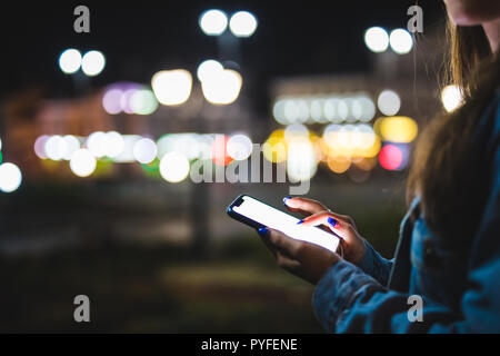 Donna puntare il dito sulla schermata vuota smartphone su sfondo bokeh luce nella notte città atmosferica, hipster utilizzando in mani pulite gadget telefono mobile Foto Stock