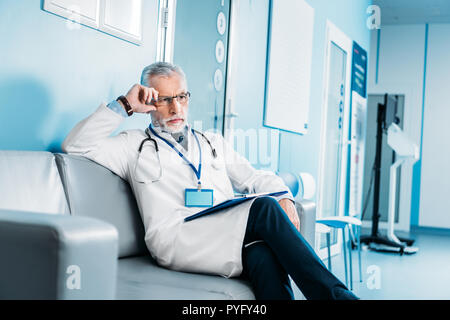 Riflessivo di mezza età medico maschio con appunti seduto sul divano nel corridoio di ospedale Foto Stock