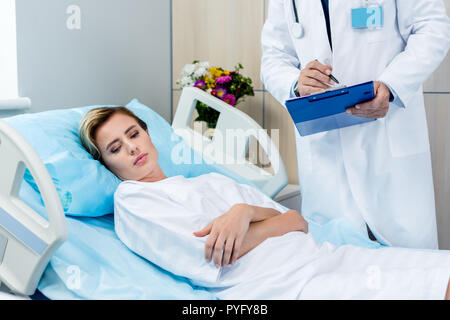 Immagine ritagliata del medico maschio con uno stetoscopio al di sopra del collo nella scrittura di appunti vicino a femmina adulta del paziente nella stanza di ospedale Foto Stock
