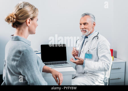 Sorridente maschio maturo medico puntando al computer portatile con schermo vuoto al paziente donna seduta vicino in ufficio Foto Stock