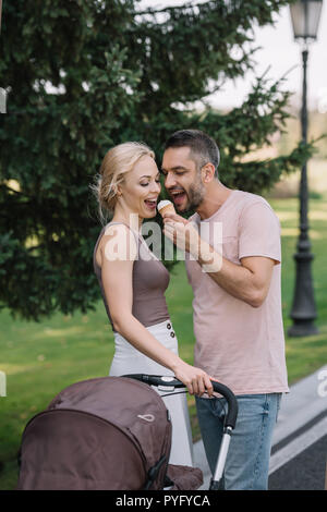 Sorridente marito moglie di alimentazione con gelato nelle vicinanze baby carrello in posizione di parcheggio Foto Stock