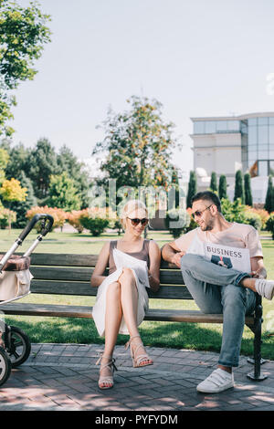 I genitori seduta sul banco vicino a baby carrello in posizione di parcheggio e la lettura di riviste e giornali di business Foto Stock