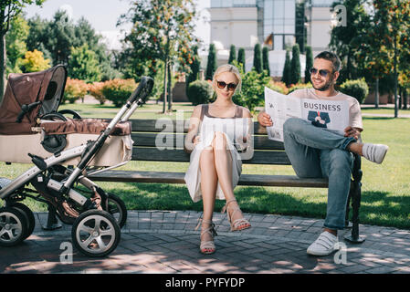 I genitori seduta sul banco vicino a baby carrello in posizione di parcheggio e la lettura di riviste e giornali Foto Stock