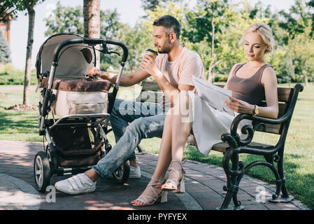 Padre di bere il caffè e toccando bambino trasporto nel parco, madre quotidiano di lettura Foto Stock