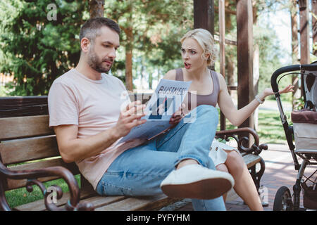 Il marito di fumare sigarette e giornale di lettura nelle vicinanze baby carrello in posizione di parcheggio, moglie a urlare contro di lui Foto Stock