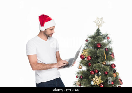 Uomo in santa claus hat utilizzando laptop vicino albero di natale isolato su bianco Foto Stock