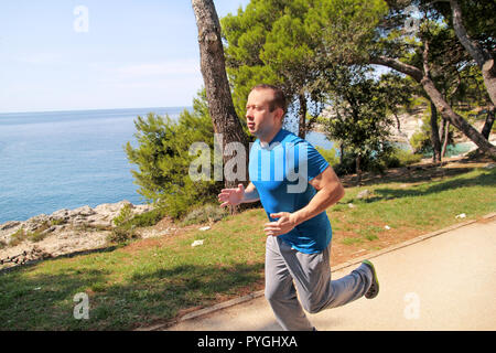 Montare il giovane uomo jogging su un acceso il sentiero lungo la riva del mare. Fitness ricreativo atleta sportswear gode di attività fisica su un giorno d'estate. Foto Stock