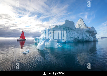 Bella rossa di barca a vela in artico accanto a un enorme iceberg che mostra la scala. Ilulissat Tourist Nature, Disko Bay, la Groenlandia. Foto Stock