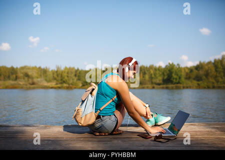 Foto di donna con le cuffie auricolari con zaino e laptop in mani seduto sulla riva del fiume sul giorno di estate Foto Stock