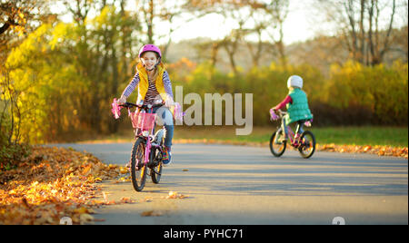 Carino piccole sorelle equitazione biciclette in un parco della città sulla soleggiata giornata autunnale. Attiva vacanza in famiglia con i bambini. Bambini indossare hemet sicurezza mentre cavalcate un modulo di interconnessione BIC Foto Stock