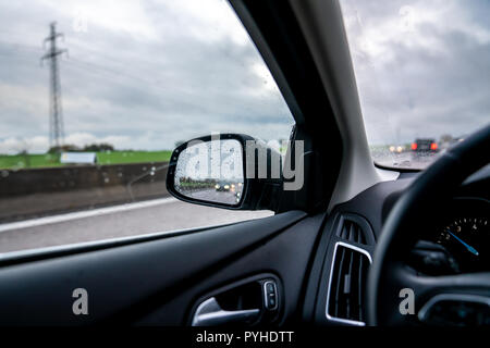 Interno del veicolo con una vista nello specchietto retrovisore dove il faro viene riflessa sotto la pioggia su una autostrada Foto Stock