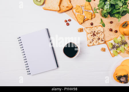 Il blocco note di vuoto e la tazza di caffè nero vicino a sandwich per colazione, vista dall'alto. Una sana toast con persimmon, kiwi, banana e dadi per spuntino mattutino su Foto Stock