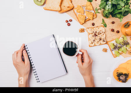 Donna di mani blank notepad e tazza di caffè forte durante il pranzo, vista dall'alto. Sana colazione toast con persimmon, kiwi, banana e dadi per Foto Stock