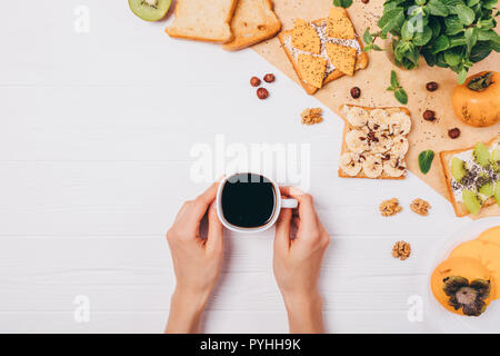 Femmina mani della coppa di ritegno di forte caffè nero vicino a sandwich per colazione, vista dall'alto. Una sana toast con persimmon, kiwi, banana e dadi per mor Foto Stock