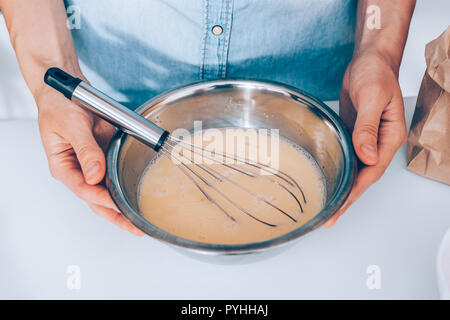 La femmina mani coppa metallica di panna uova con frusta accanto al sacchetto di farina bianca sul tavolo da cucina, vista dall'alto. Close-up donna fatto impasto per pan Foto Stock