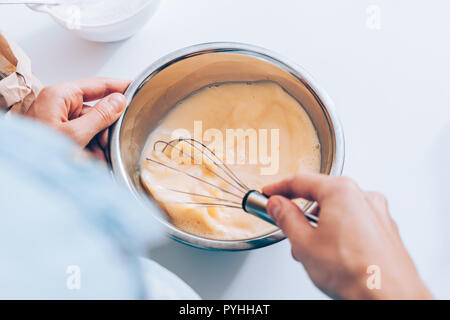 Close-up della femmina mani ciotola sbattere l'impasto liquido di latte e uova sul tavolo bianco. Immagine candidi donna mescolando la pastella per i pancake. Foto Stock