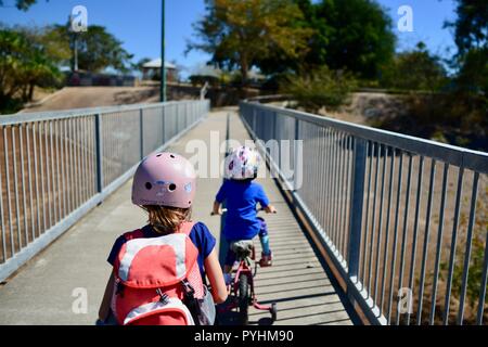 I bambini piccoli in sella a una moto e scooter attraverso un ponte pedonale, Aplin's weir a Ross River, Townsville, QLD, Australia Foto Stock