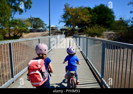 I bambini piccoli in sella a una moto e scooter attraverso un ponte pedonale, Aplin's weir a Ross River, Townsville, QLD, Australia Foto Stock