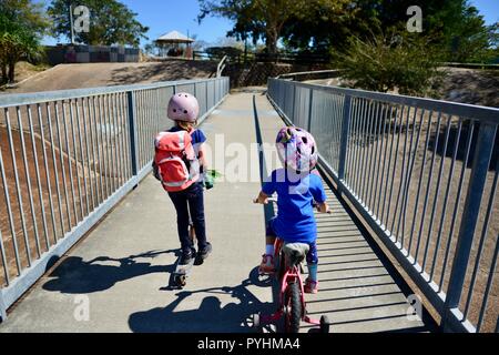 I bambini piccoli in sella a una moto e scooter attraverso un ponte pedonale, Aplin's weir a Ross River, Townsville, QLD, Australia Foto Stock