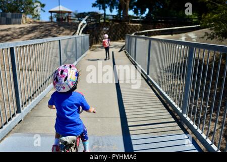 I bambini piccoli in sella a una moto e scooter attraverso un ponte pedonale, Aplin's weir a Ross River, Townsville, QLD, Australia Foto Stock