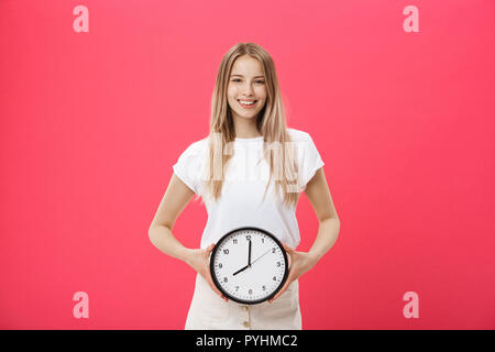 Ritratto di un entusiasta giovane ragazza vestita di bianco t-shirt puntando alla sveglia e guardando la telecamera isolate su sfondo rosa Foto Stock