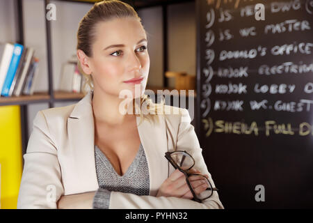 Ritratto di una giovane donna bellissima con gli occhiali in mani in piedi in classe e grave di insegnante di istituto di istruzione superiore, intelligente e di successo Foto Stock