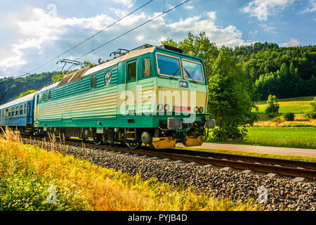 Un verde locomotiva elettrica passando la campagna ceca. Un treno che corre attraverso la valle verde. Il trasporto ferroviario in Repubblica Ceca. Una giornata di sole Foto Stock