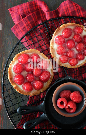 Rosso lampone frollini crostate con vanille crema pasticcera e vetrata di lamponi freschi sul raffreddamento per rack in legno marrone scuro, vista dall'alto Foto Stock