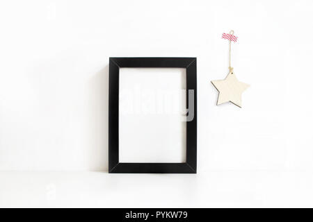 Verticale nera vuota telaio in legno mockup sul tavolo bianco. La stella di legno decorazione appesa al muro. Stile femminile stock photography. Home decor. Natale concetto d'inverno. Foto Stock