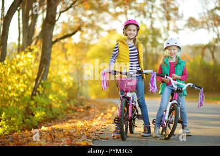 Carino piccole sorelle equitazione biciclette in un parco della città sulla soleggiata giornata autunnale. Attiva vacanza in famiglia con i bambini. Bambini indossare hemet sicurezza mentre cavalcate un modulo di interconnessione BIC Foto Stock