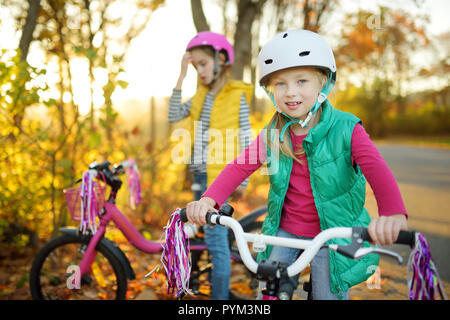 Carino piccole sorelle equitazione biciclette in un parco della città sulla soleggiata giornata autunnale. Attiva vacanza in famiglia con i bambini. Bambini indossare hemet sicurezza mentre cavalcate un modulo di interconnessione BIC Foto Stock