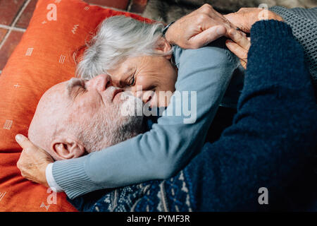 Donna sorridente dormire nelle braccia di suo marito tenendo la sua testa. Felice coppia senior di dormire sul pavimento che abbraccia ogni altra in una fredda notte. Foto Stock