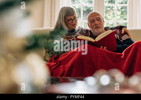 Coppia senior avente un tempo di rilassarsi leggendo un libro seduti a casa insieme. Il vecchio uomo e donna seduta insieme su un divano coperto di una coperta. Foto Stock
