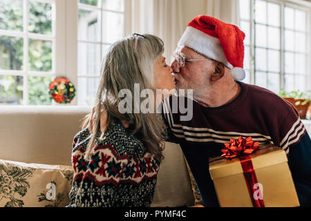 Senior uomo in santa cap baciando la moglie tenendo un regalo di Natale. Coppia senior festeggiano il Natale e il messaggio di saluto di ogni altro a casa. Foto Stock