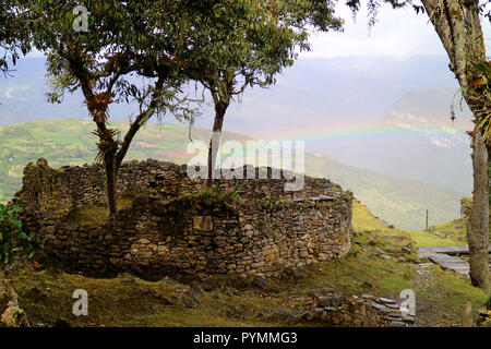 Alberi che crescono all'interno di casa arrotondati rovine di Kuelap mountaintop cittadella con l'arcobaleno della bassa collina in background, nord del Perù Foto Stock