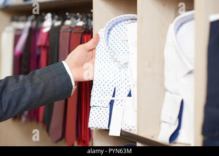 Stretta di mano d'uomo tenendo la camicia bianca in blu pattern. Shelfe con maschio camicie e appendiabiti con fascette colorate sullo sfondo. Raccolto di abbigliamento maschile e accesorize. Concetto di shopping e della moda. Foto Stock