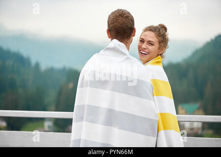 Coppia giovane indossare costumi da bagno nel confezionamento in striped asciugamani, permanente sulla terrazza e guardare le montagne di nebbia. Curly bella ragazza, abbracciando il suo fidanzato, sorridente e guardando la fotocamera. Foto Stock